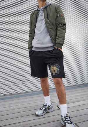 Uomo in piedi con le mani nelle tasche, indossa una giacca bomber verde, felpa grigia con cappuccio, pantaloncini neri con stampa, calzini bianchi e sneakers grigie su un ponte di legno.