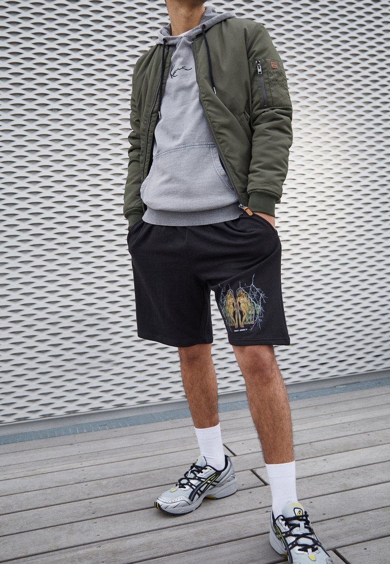 Un homme se tient debout, les mains dans les poches, portant une veste bomber verte, un sweat à capuche gris, un short graphique noir, des chaussettes blanches et des baskets grises sur une terrasse en bois.