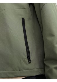 Veste vert olive avec une texture lisse, comportant une poche zippée noire sur le côté. Dans l'ensemble, un design simple et pratique.