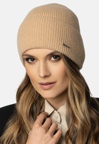 Beige gebreide beanie met ribbelstructuur en gestikte logodetail. Zacht materiaal en nauwsluitend ontwerp.