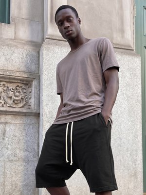 Jeune homme portant un t-shirt marron clair et un short noir avec des cordons blancs, debout contre un mur en pierre avec une décoration sculptée.
