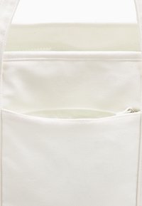 Bijela platnena torba s teksturiranom površinom ima zip džep sprijeda i gornju ručku za nošenje. Jednostavan i funkcionalan dizajn.