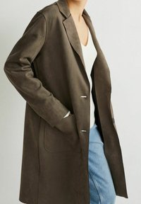 Abrigo de ante marrón con una silueta ajustada, solapas en pico, dos bolsillos laterales y detalles de hardware en tono plateado. Llevado sobre una blusa blanca y unos vaqueros.