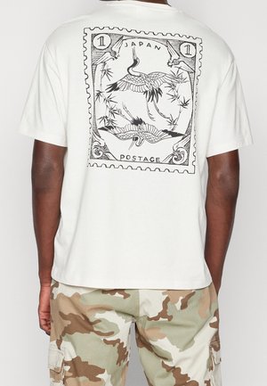 Biały bawełniany t-shirt z czarnym nadrukiem przedstawiającym żurawie i bambus, otoczony dekoracyjną ramką, na plecach.