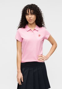 Ροζ ανοιχτό πουκάμισο πόλο με γιακά, κοντά μανίκια και κεντημένο κόκκινο λογότυπο στο στήθος. Συνδυασμένο με σκούρο πλισέ φούστα.