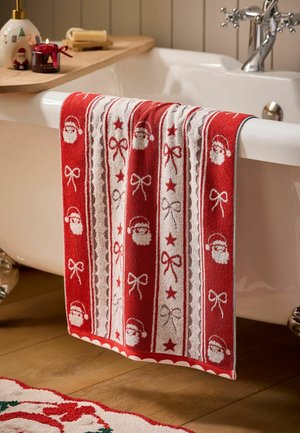 Serviette rouge et blanche avec Père Noël, étoiles et nœuds suspendue à une baignoire blanche avec un plateau en bois tenant une bougie et une bouteille de lotion.