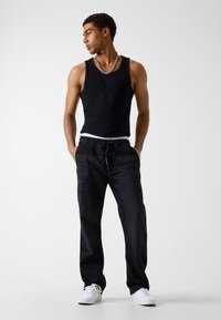 Débardeur noir, pantalon ample noir, ceinture blanche visible et baskets blanches. Le modèle est debout, les mains dans les poches, regardant sur le côté.
