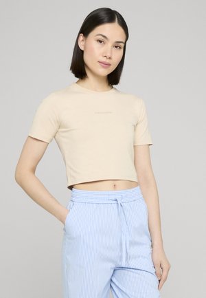 Ung kvinde med glat mørkt hår bærer en beige croppet t-shirt og blå og hvide stripede bukser med hænderne i lommerne.