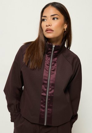 Femme aux cheveux bruns lisses portant une veste zippée marron foncé avec des garnitures brillantes et des boucles d'oreilles anneaux dorées, regardant sur le côté
