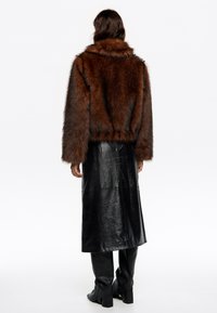 Brązowa kurtka z sztucznego futra z wysokim kołnierzem, nałożona na czarną skórzaną długą spódnicę, w połączeniu z czarnymi kozakami do kolan.