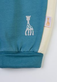 Tessuto teal con una texture a coste e polsini elasticizzati. Presenta un design di giraffa ricamato e un piccolo etichetta logo in rosa chiaro sul lato crema.