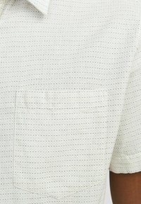 Camisa de manga corta en tejido beige claro con un patrón texturizado y puntos en azul marino. Cuenta con un bolsillo en el pecho y un dobladillo recto.