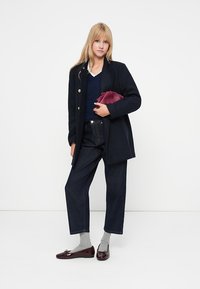 Mornarski plašč s pozlačenimi gumbi, nošen čez mornarsko pulover, v kombinaciji s temnim jeansom in burgundskimi loaferji; drži burgundsko torbico.