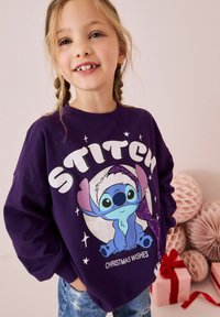 Lilla dress, millel on Stitchi kujutis sädelevate aktsentidega. Tekst ütleb "STITCH" ja "JÕULU SOOVID." Näha on denimid.