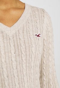 Pull beige en maille torsadée à col en V, manches longues, avec un petit logo oiseau rouge brodé sur la poitrine gauche.