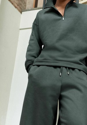 Personne portant un sweat-shirt à fermeture éclair vert foncé et un pantalon de survêtement assorti, les mains dans les poches, debout à l'intérieur près d'un mur en briques.