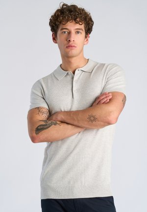 Joven con cabello rizado y tatuajes en ambos brazos, vistiendo una camisa polo de manga corta color gris claro, con los brazos cruzados y expresión neutral.