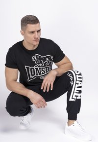 Črna majica Lonsdale z belim grafičnim motivom leva; črne trenirke z belim logotipom; model nosi bele superge; minimalističen dizajn, atletski kroj.