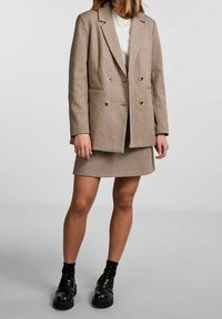 Kobieta w beżowej dwurzędowej marynarce i dopasowanej spódnicy, w białej bluzce, czarnych skarpetkach w kropki i czarnych masywnych butach, stojąca na szarym tle.