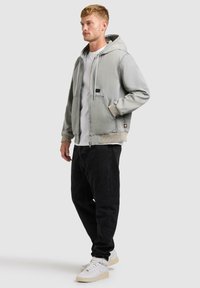 Jeune homme debout portant une veste à capuche gris clair, un t-shirt blanc, un pantalon noir et des baskets blanches sur un fond uni.