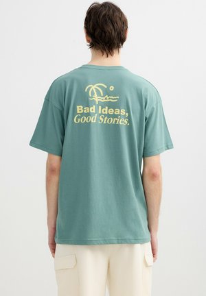Persona que lleva una camiseta verde azulado con un gráfico amarillo de una palmera y el texto "Malas ideas, buenas historias." en la parte trasera.