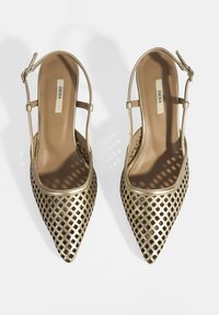 Talons aiguilles en maille dorée avec des découpes, une bride de cheville et une semelle intérieure beige lisse. La surface texturée crée un motif en treillis.