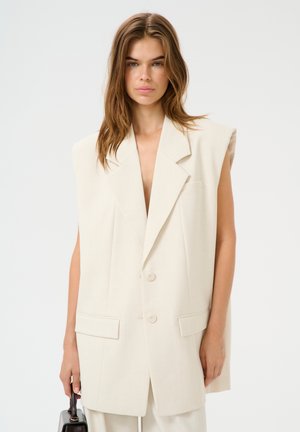 Beige übergroßer ärmelloser Blazer mit großen geknöpften Revers, zwei vorderen Taschen und Doppelknopfverschluss. Glatte Textur und taillierte Passform.