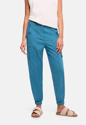 Mujer que lleva pantalones jogger cargo azules con puños elásticos y sandalias beige, combinados con un top blanco sin mangas.