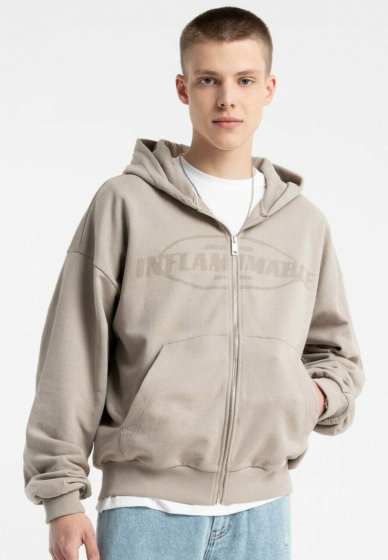 Beżowa bluza z kapturem na zamek z dużym napisem "INFLAMMABLE". Posiada regulowany kaptur, boczne kieszenie i ściągacze na mankietach. Wykonana z miękkiego materiału.