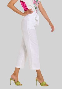 Weißer Leinenhose mit hoher Taille und Schleifen-Gürtel, kombiniert mit grünen geflochtenen Sandalen mit Absatz, vor einem grauen Hintergrund gestylt.