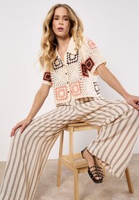 Top all'uncinetto con motivo a quadri in arancione e marrone. Pantaloni beige a righe larghe. Sandali neri con borchie. Modella seduta su uno sgabello di legno.