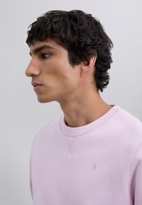 Joven con cabello corto y rizado de color oscuro, vistiendo un sudadera de cuello redondo de color rosa pastel, mirando hacia la izquierda contra un fondo claro y liso.