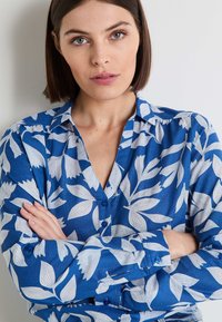 Camisa azul de botones con patrones florales blancos, mangas largas con puños abotonados y un corte relajado. Textura de tela suave.