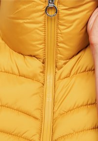 Żółta kamizelka typu puffer z pikowaną teksturą, z metalowym zamkiem błyskawicznym oraz logiem umieszczonym wzdłuż linii zamka. Zbliżenie na tkaninę i elementy metalowe.