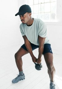 Athletischer Mann in einem hellblauen feuchtigkeitsableitenden Shirt und marineblauen Shorts, der eine schwarze Kettlebell hebt. Er trägt graue Sneaker und eine schwarze Kappe.