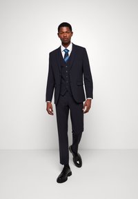 Marine drie-delige pak met een slim fit, voorzien van een notch revers, vijf knopen op de gilet en een gestreepte blauwe stropdas. Zwarte veterschoenen.