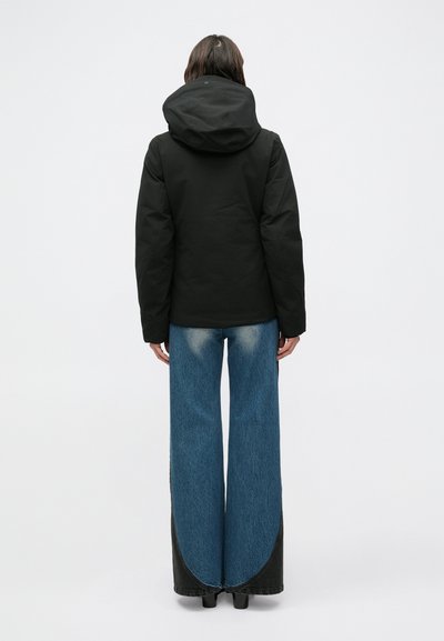 Crna izolirana jakna s velikim kapuljačom, uparena s širokim plavim trapericama s blagim izblijedjelim detaljima i crnim naglaskom na rubu.