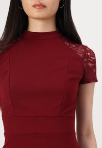 Rochie burgundy cu mâneci scurte din dantelă, guler înalt și un corset ajustat. Textură de material neted cu cusături structurate și un aspect elegant.