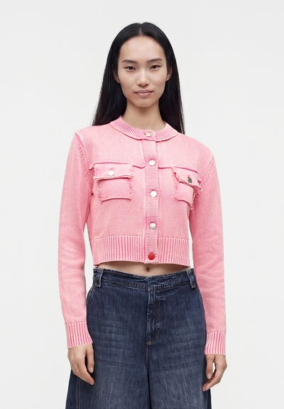 Jeune femme portant un cardigan court rose avec des boutons et deux poches devant, associé à un jean bleu foncé taille haute.