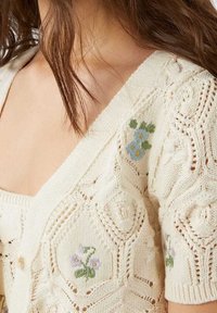 Off-white knitted cardigan featuring a textured pattern, short sleeves, and floral embroidery in blue and purple on the chest.