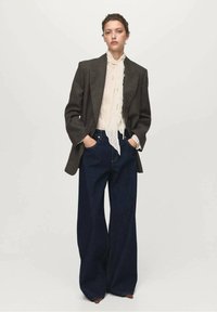 Blazer de tweed marrón sobre una blusa crema con volantes, combinado con jeans de mezclilla oscuros de pierna ancha. Cintura alta notable y silueta alargada.