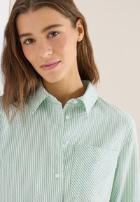 Jeune femme aux cheveux châtain clair portant une chemise à boutons à rayures verticales vert clair et blanc avec col et poche poitrine.