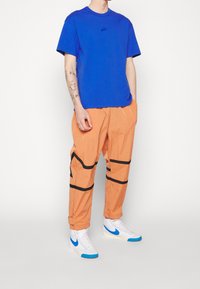 Modré tričko Nike, krátké rukávy, s malým logem. Oranžové lehké kalhoty, elastický pas, černé dekorativní pruhy, v kombinaci s bílými a modrými botami.