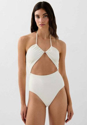 Mujer con cabello oscuro vistiendo un bañador blanco con cuello halter y una abertura en forma de ojo de cerradura sobre el abdomen, de pie frente a un fondo liso.