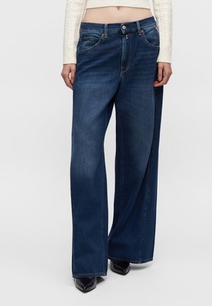 Weit geschnittene dunkelblaue Jeans, kombiniert mit einem weißen kurzen Pullover und schwarzen spitzen Schuhen, von der Taille bis zu den Füßen gezeigt.