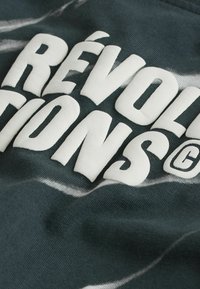 Gros plan sur un tissu foncé avec de grandes lettres blanches en relief formant le mot "RÉVOLUTIONS" et un petit symbole "G" cerclé.