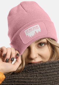 Pink knit beanie with ribbed texture, featuring a rectangular patch with white forest graphics and text. Warm and fitted design.