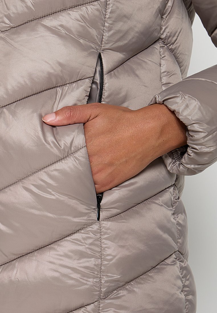 Mano inserita nella tasca con zip di un giubbotto imbottito trapuntato taupe lucido, con polsino elastico sulla manica.