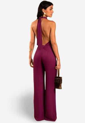 Une combinaison salopette prune avec un dos ouvert et des jambes évasées, présentant un col noué, des détails de couture minimaux et une texture lisse. Compléments avec un petit sac.