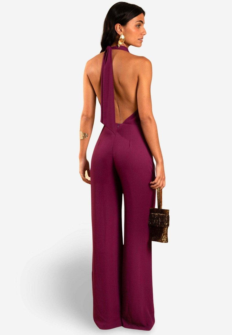 Mono de tirantes color ciruela con espalda descubierta y piernas acampanadas, que cuenta con un cuello atado, detalles de costura mínimos y una textura suave. Se complementa con un bolso pequeño.